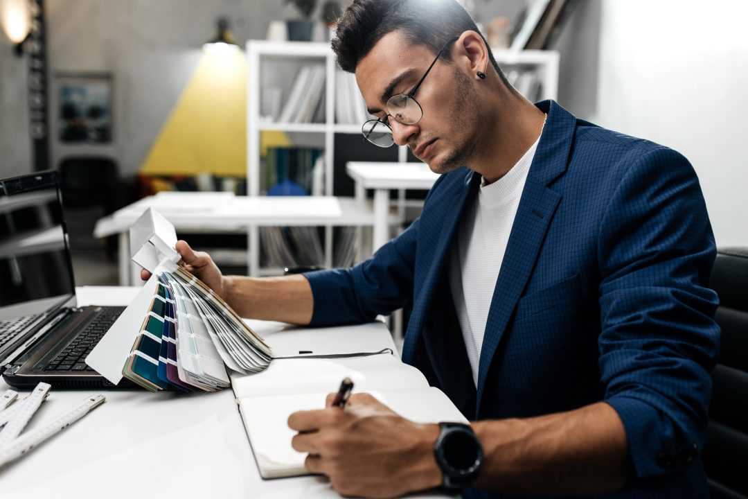 The architect in glasses and in a blue jacket is working with catalog of colors on the desk in the