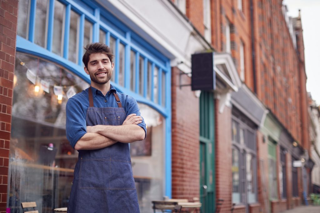 Portrait Of Male Small Business Owner Wearing Apron Standing Outside Shop On Local High Street